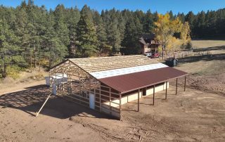 A partially constructed barn with a brown roof and unfinished walls stands on dirt ground, surrounded by trees and near a house.