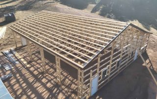 A partially constructed wooden barn with an open framework and a slanted roof, surrounded by a dirt area and scattered building materials.