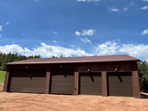 A large brown metal garage shop in Douglas County, featuring four closed roll-up doors, set on a dirt lot with trees and blue sky in the background.
