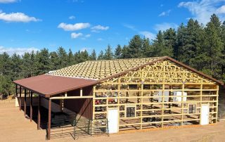 Partially constructed wooden pole barn with a metal roof on one section, surrounded by dirt ground and trees in the background—ideal for those looking to shop Douglas County for rural building projects.