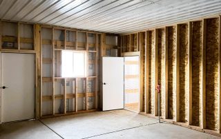 Interior of an unfinished room with exposed wooden wall studs, a window, two white doors, and a concrete floor. Ceiling is covered with metal panels—perfect inspiration if you’re looking to shop Douglas County for renovation ideas.