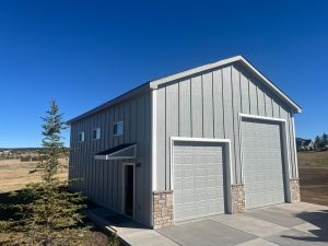 Media - Alt Text A modern two-story gray garage with vertical siding, two large garage doors, stone accents, and a small porch, set against a clear blue sky and open landscape.