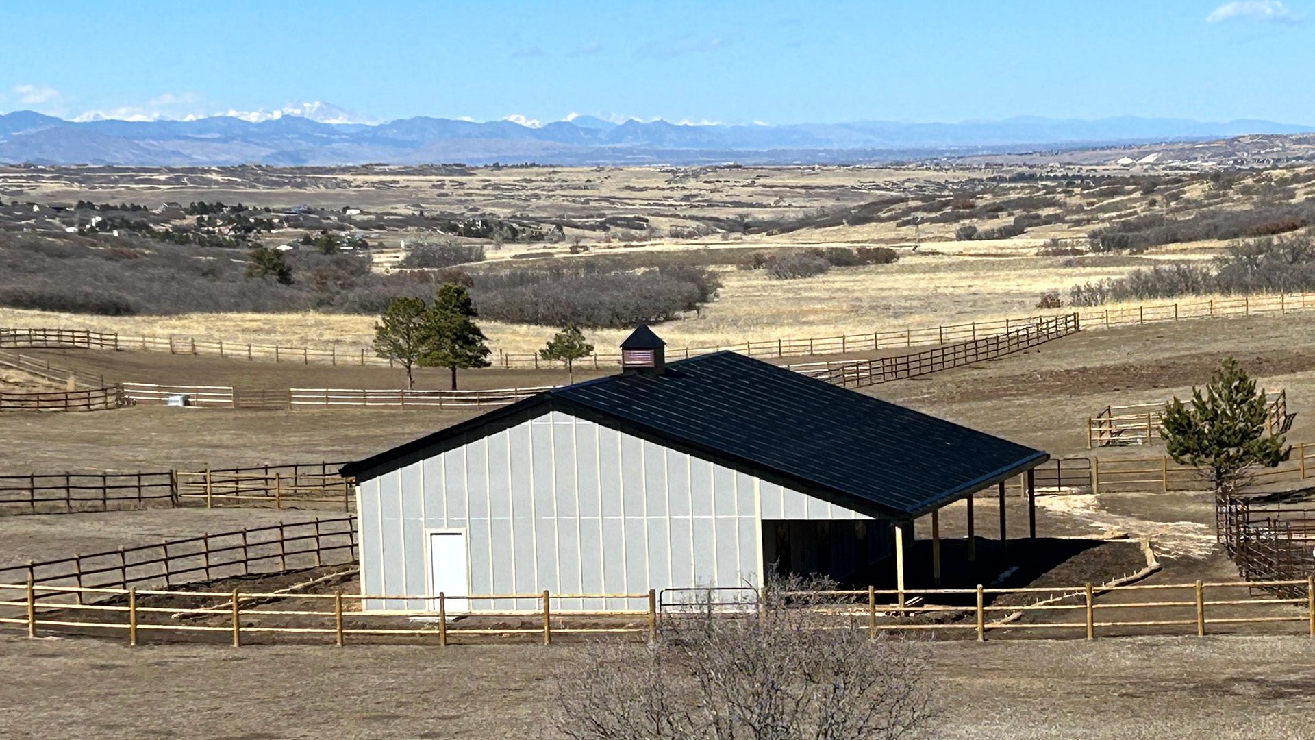 A custom horse barn with a black roof is surrounded by fenced paddocks in a dry, rural landscape, set against distant mountains.