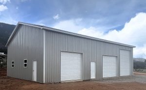 A large, gray metal building with two white garage doors and two white entry doors, set on a dirt lot under a partly cloudy sky.