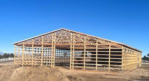 A wooden frame of a large barn structure under construction stands on a dirt lot under a clear blue sky.