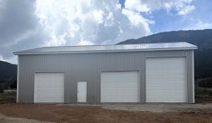 A large metal building with a metal roof, three white garage doors of varying sizes, and a single white entry door, set against a cloudy sky and mountain backdrop.