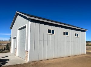 A light gray detached garage with two large white doors and three small rectangular windows, situated on a flat dirt and concrete surface under a clear blue sky.