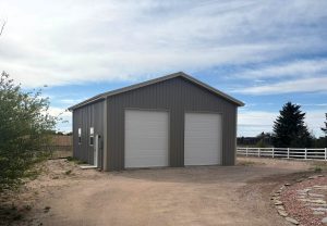 A metal building with two large white garage doors sits on a dirt lot, surrounded by a fence and some trees under a partly cloudy sky.