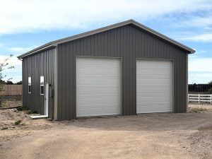 A gray metal garage with two large white roll-up doors and a side entrance door, situated on a dirt driveway under a partly cloudy sky.