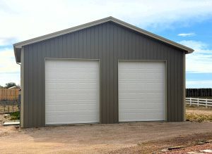 A gray metal building with two closed white garage doors on a gravel driveway, set against a partly cloudy sky.
