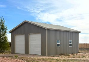 A metal building with two large white garage doors and two small windows, situated on a dirt lot with a wooden fence in the background.