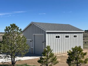 A gray wooden shed with a gabled roof, three small windows, and a closed garage door, set on a concrete pad amid sparse trees under a clear blue sky.