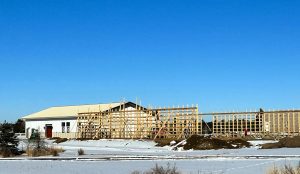 A partially constructed wooden structure stands beside a completed white building with a metal roof on a snowy landscape under a clear blue sky.