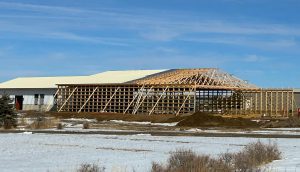 A large wooden-framed building under construction stands on a snowy, grassy field beneath a blue sky.