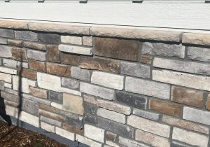 A section of a building exterior featuring a pattern of rectangular stone bricks in various shades of gray, brown, and beige under a white siding.