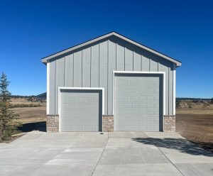 A gray building with two garage doors of different sizes, set on a concrete driveway with a clear blue sky in the background.