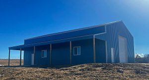 A large blue metal building with a covered porch and several windows sits on a dry, grassy plot under a clear blue sky.