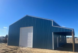 A large blue metal building with a white garage door and a covered side entry, situated on a dirt lot under a clear blue sky.