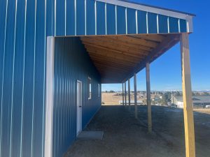 A blue metal building with a wooden overhang supported by posts, overlooking an open dirt area and distant houses under a clear sky.