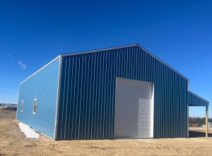 A blue metal building with a large white garage door and small windows stands on a dirt lot under a clear blue sky.