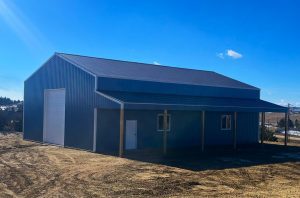 A blue metal building with a large garage door, a smaller entry door, and windows sits on a dirt lot under a clear blue sky.