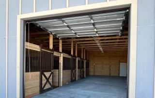Open garage-style door reveals the interior of a barn with horse stalls on the left and a concrete floor. Snow is visible on the ground outside.