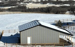 A gray shed with a slanted roof stands in a snow-covered fenced area, with tracks visible in the snow and hills in the background.