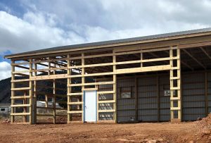 A partially constructed wooden frame building with a metal roof and siding, featuring a white door and windows, on a dirt lot under a partly cloudy sky.