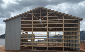 Partially constructed wooden frame of a large shed or barn with some metal siding, set on a dirt lot with mountains in the background under a cloudy sky.