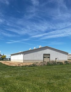 A large gray metal barn with a closed garage door sits on a grassy lot under a blue sky with wispy clouds.