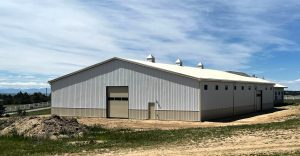 A large white metal barn with a closed garage door stands on a grassy lot under a blue sky with clouds.