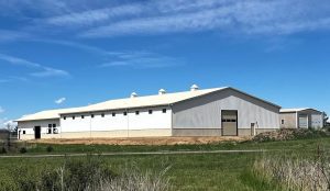 Large white industrial warehouse building with a light gray roof, set on a grassy lot under a blue sky with scattered clouds.