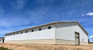 Large white industrial warehouse building with small windows near the roof and a large garage door, set against a blue sky with light clouds.