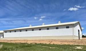 A large white metal building with multiple small windows and rooftop vents sits on a grassy lot under a blue sky with scattered clouds.