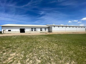 A large white metal barn with several windows and doors sits on dirt and sparse grass under a blue sky with scattered clouds.