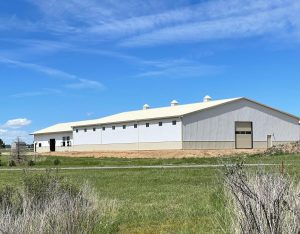 Large white metal barn with a light-colored roof stands on a grassy lot under a blue sky with scattered clouds.