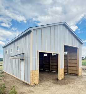 A partially finished light blue metal building with two large open garage-style doors, small windows, and plywood panels at the base, set on dirt under a partly cloudy sky.