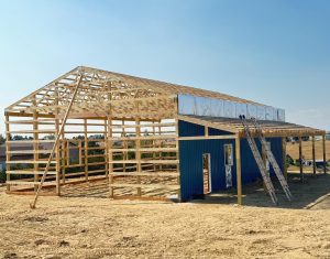 Partially constructed blue metal building with exposed wooden roof trusses, a large ladder leaning against the side, and dry ground in the foreground.