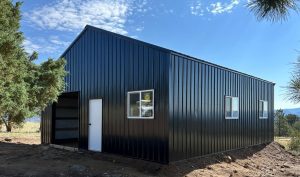 A black metal building with a white door and several windows stands on a dirt lot surrounded by trees under a partly cloudy sky.