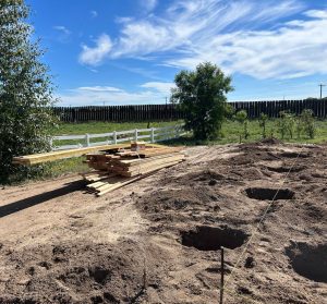 A stack of wooden planks is placed on the dirt beside several dug holes and a string line, with a white fence and trees in the background under a blue sky.