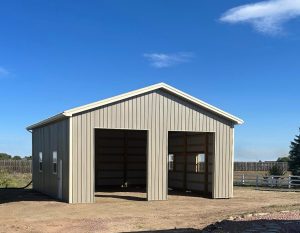A beige metal barn with two large open garage-style doors stands on a dirt lot under a clear blue sky.