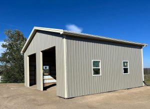 A beige metal building with two large open garage doors and two small windows, standing on a dirt lot under a clear blue sky.