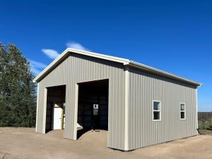 A light gray metal building with two large open garage doors and two small side windows stands on a dirt lot under a clear blue sky.