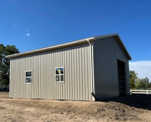 A beige metal building with vertical siding, two windows, and a large open doorway stands on bare ground under a clear blue sky.