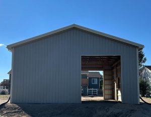 A gray metal barn with an open front facing the viewer, revealing its unfinished interior and houses visible in the background.