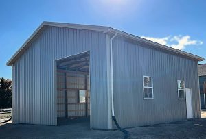 A large, gray metal-sided building with an open garage door, two windows, and a downspout, standing under a clear blue sky.