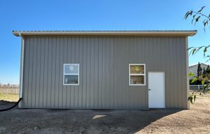 A gray metal building with two windows and a white door on one side, set on bare ground under a clear blue sky.