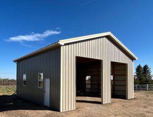 A tan metal building with two large open garage bays, a small white side door, and windows, set on dirt with a clear blue sky in the background.