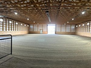 Indoor riding arena with sand footing, wooden walls, exposed roof trusses, overhead lights, and a partially open garage-style door at the far end.