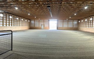 Indoor riding arena with sand footing, wooden walls, exposed roof trusses, overhead lights, and a partially open garage-style door at the far end.